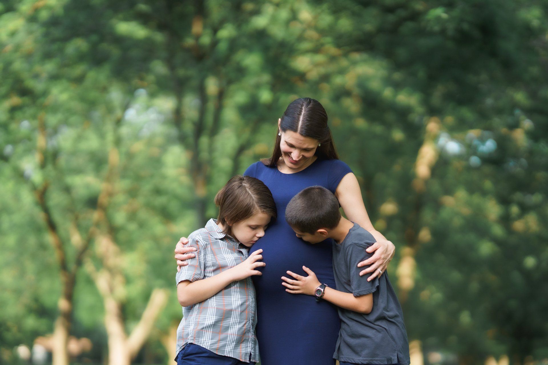 A pregnant woman is being hugged by two boys in a park.