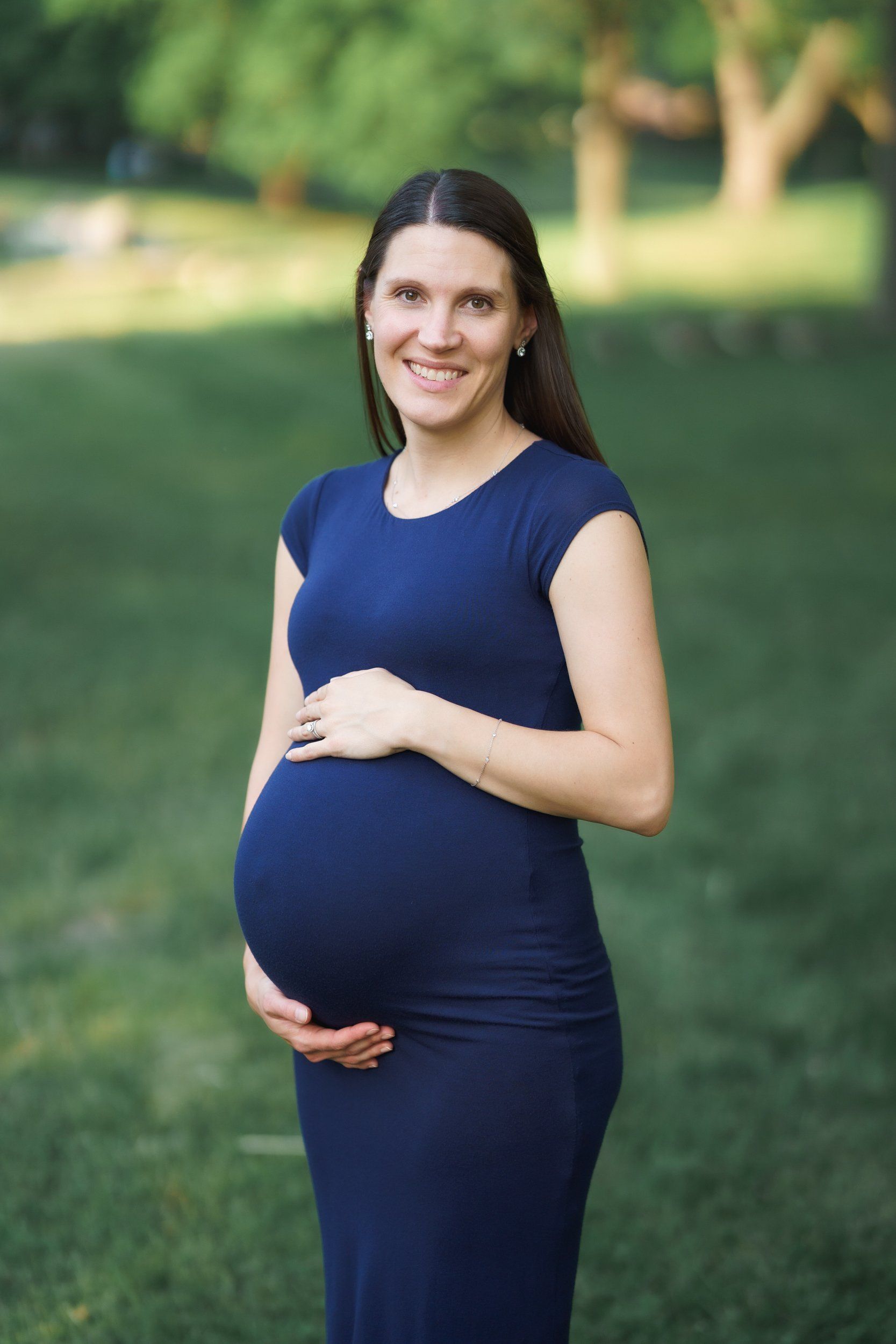 A pregnant woman in a blue dress is holding her belly in a park.