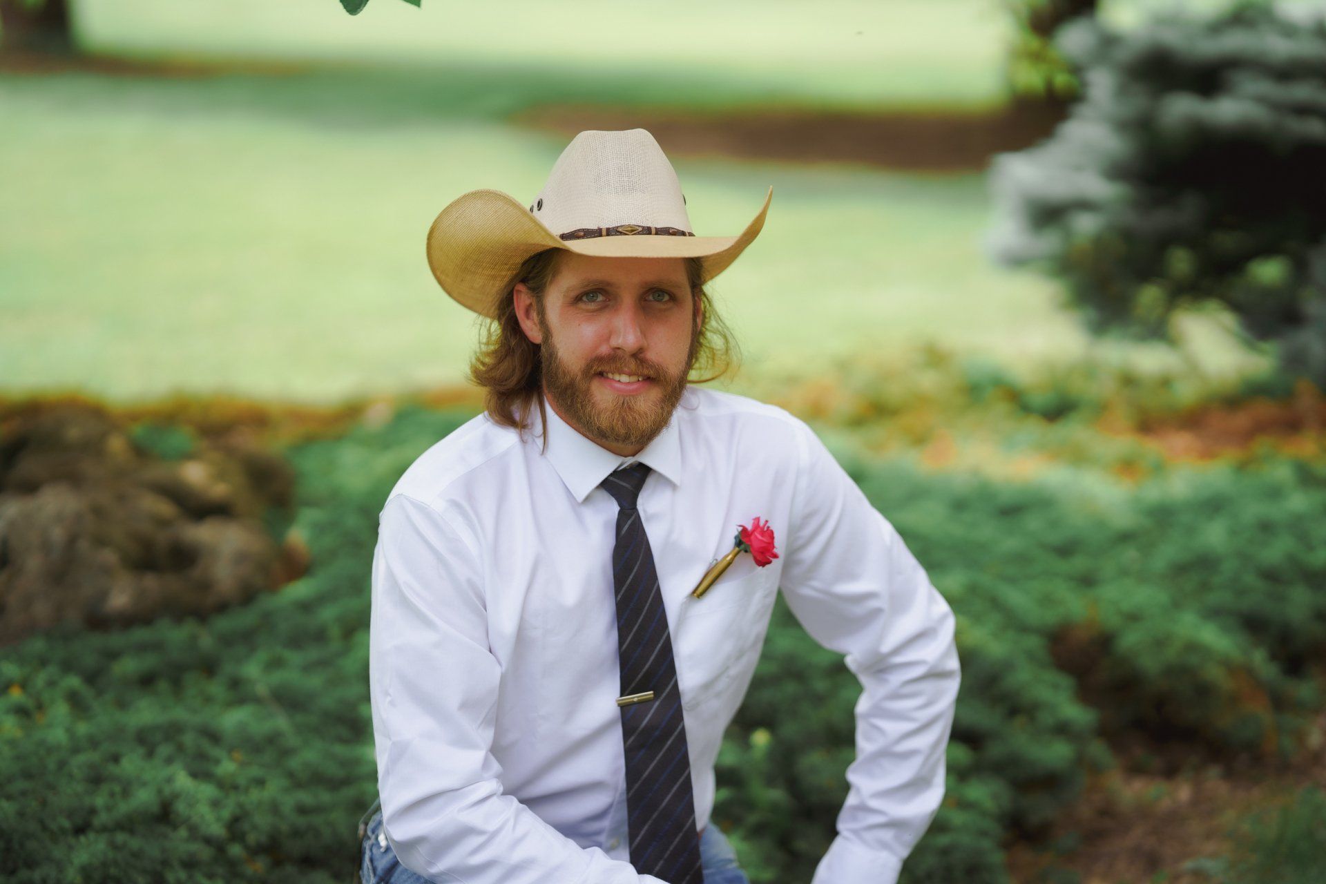 A man wearing a cowboy hat and tie is sitting in the grass.
