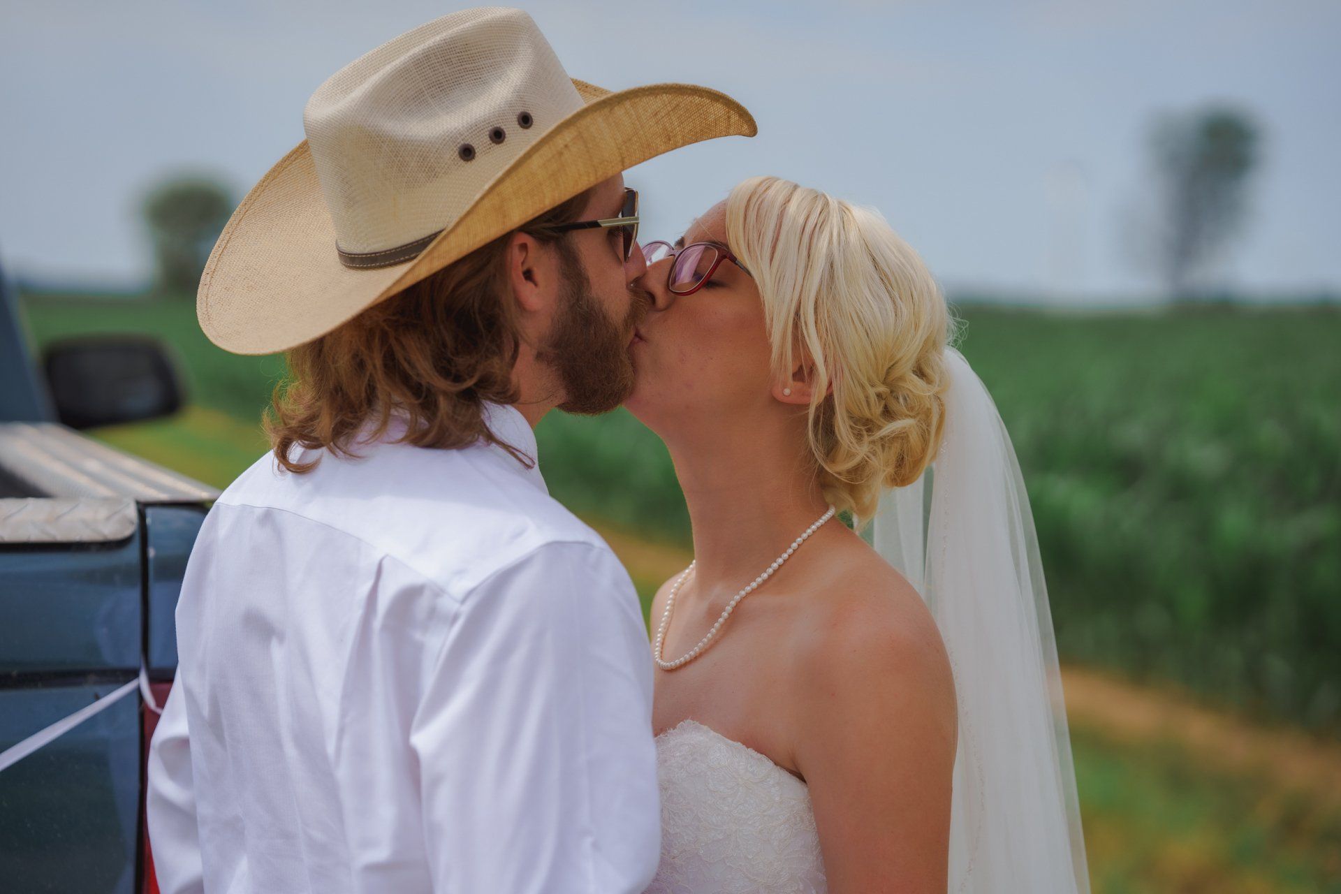 A bride and groom kissing in front of a truck . the bride is wearing a cowboy hat.