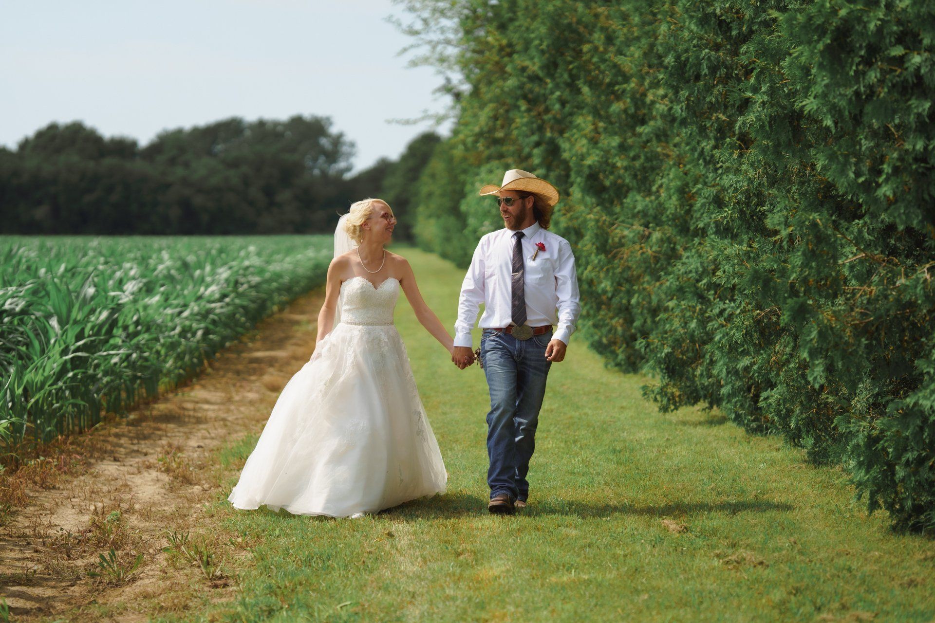 A bride and groom are walking down a path in a field holding hands.