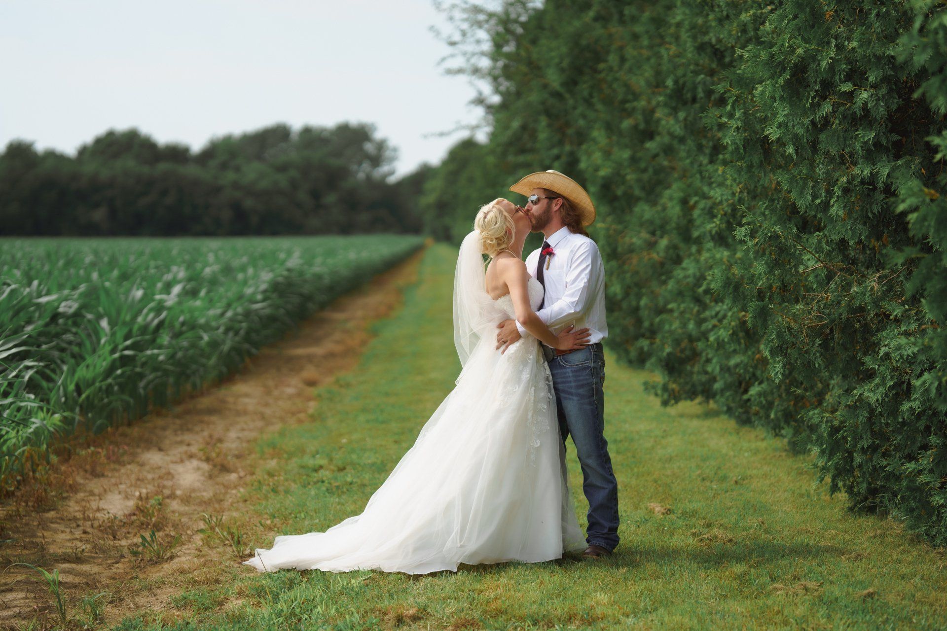 A bride and groom are kissing in a field . the bride is wearing a cowboy hat.
