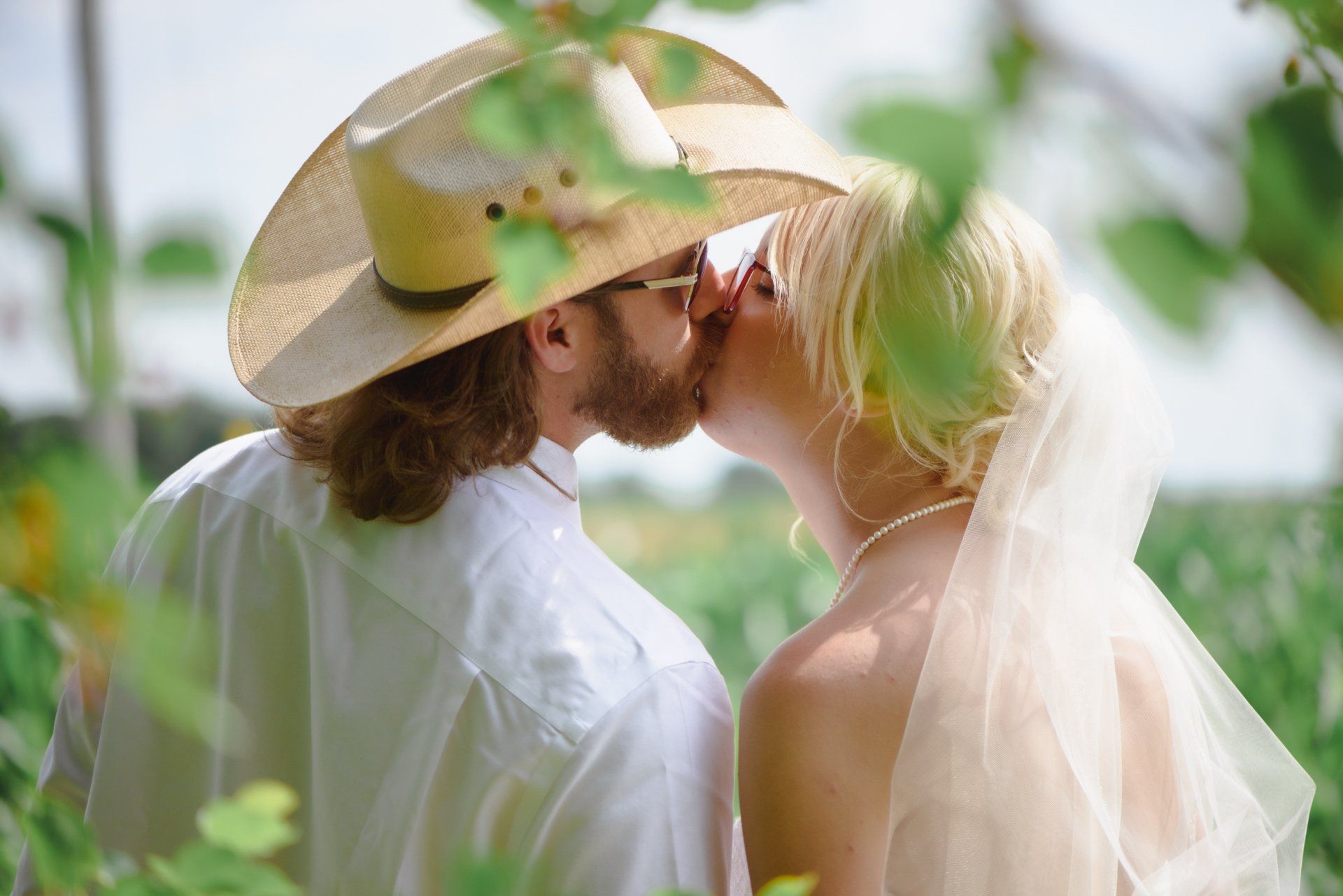 A bride and groom are kissing in a field while the bride is wearing a cowboy hat.