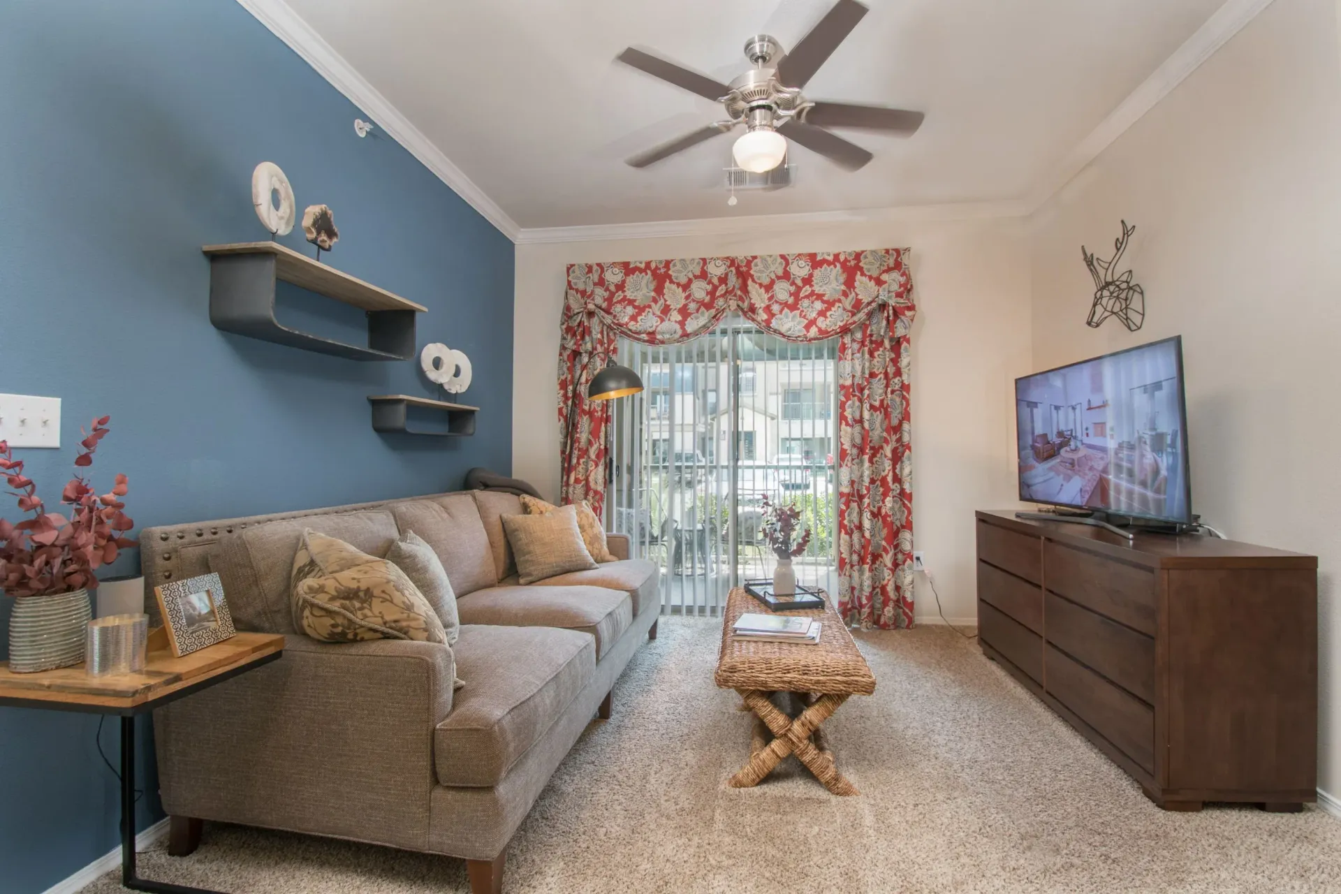 Apartment living room with a blue accent wall, tan sofa, red floral curtains, ceiling fan, and TV.