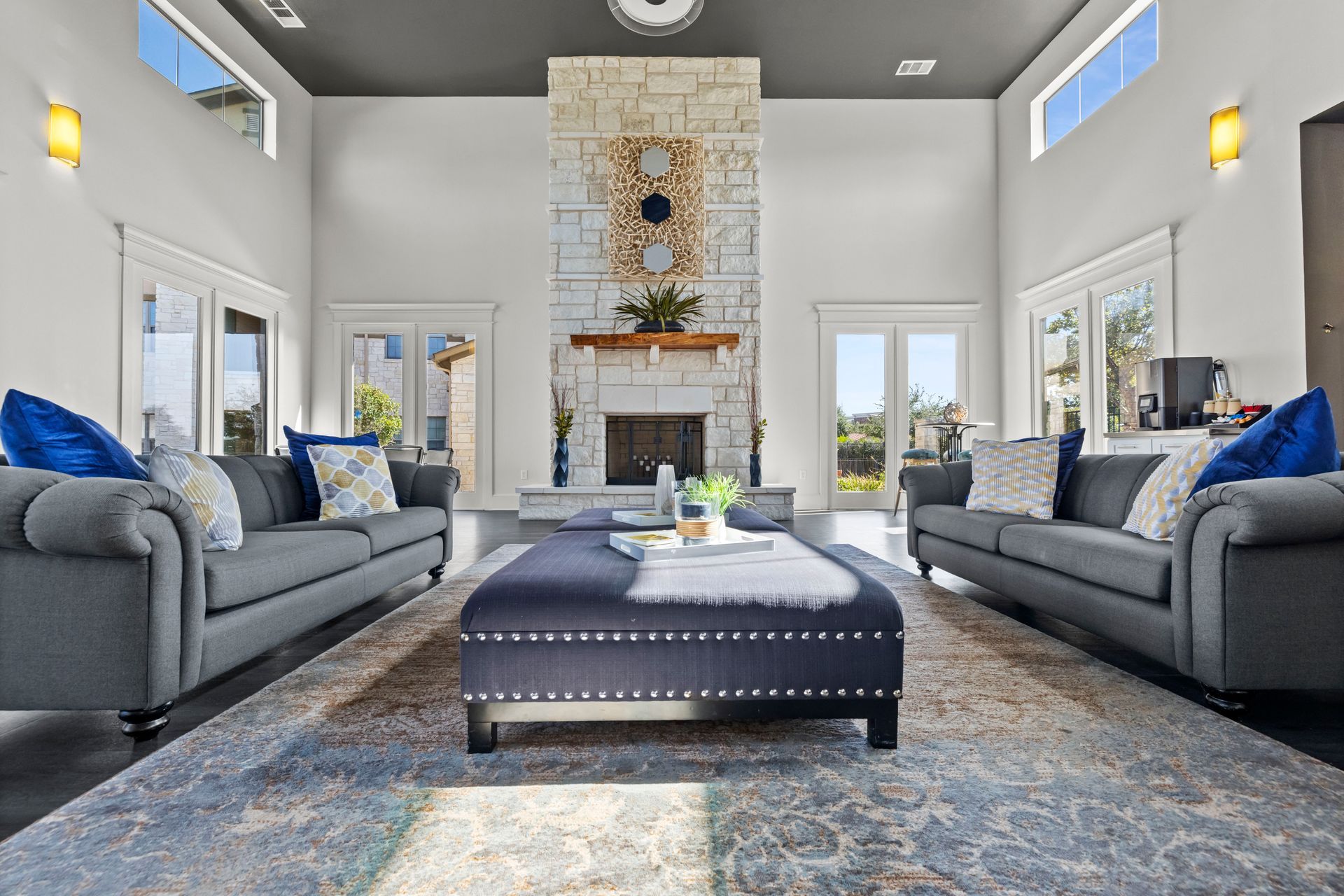 A bright living room with two gray sofas, a large navy ottoman, and a tall stone fireplace under a dark ceiling.