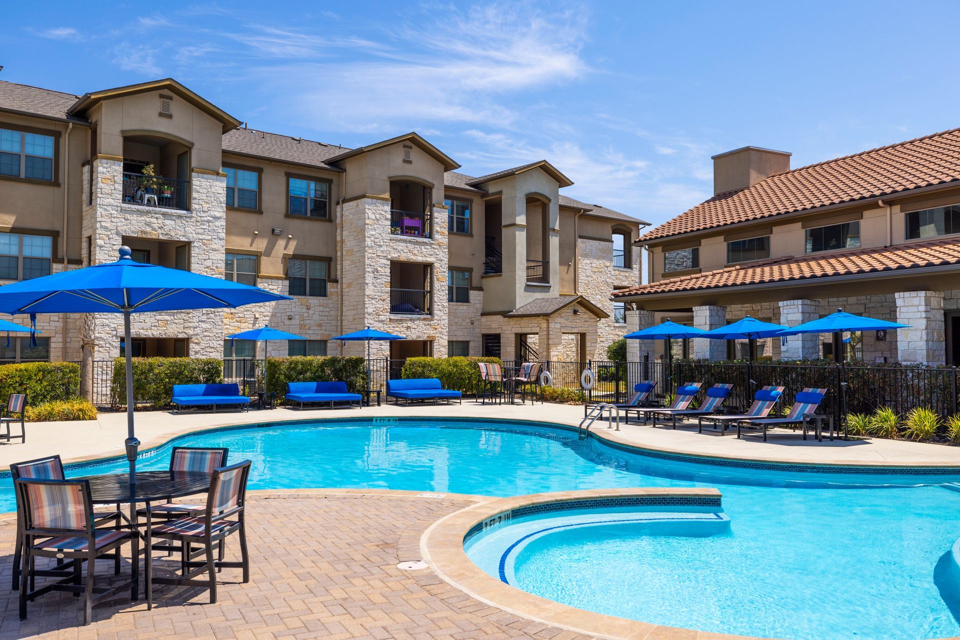 A bright, sunlit apartment swimming pool area with blue umbrellas, lounge chairs, and tan stone-facade buildings.