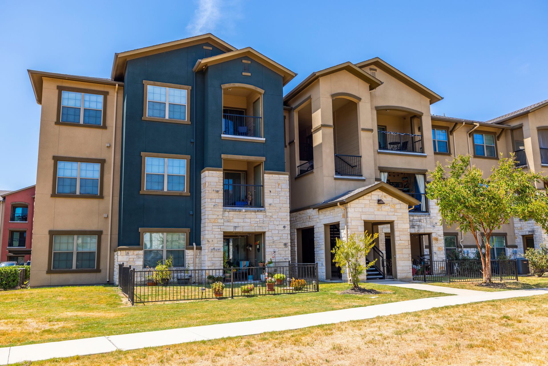 A three-story apartment complex featuring blue and tan stucco siding with stone accents under a clear blue sky.
