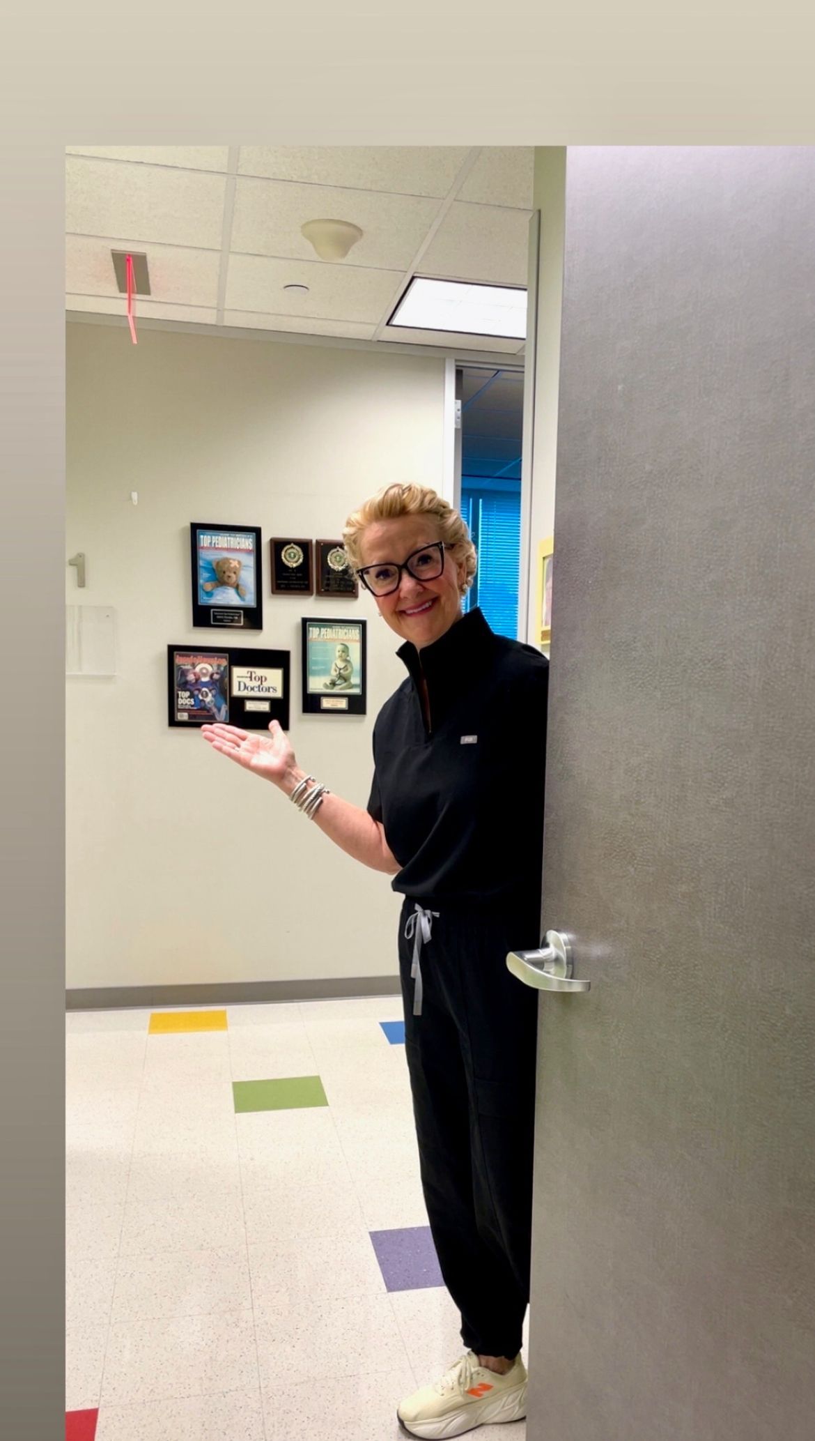 Woman in black scrubs gestures from a doorway in a hallway with colorful floor tiles and framed pictures.