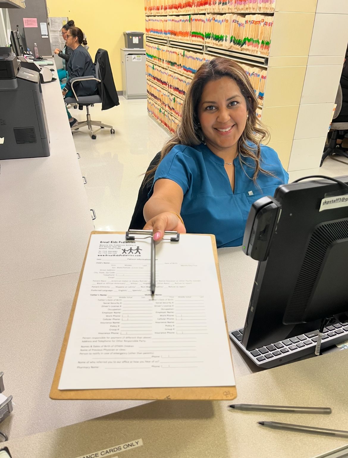 Woman in an office with a computer screen in front of her and medical files in the background.
