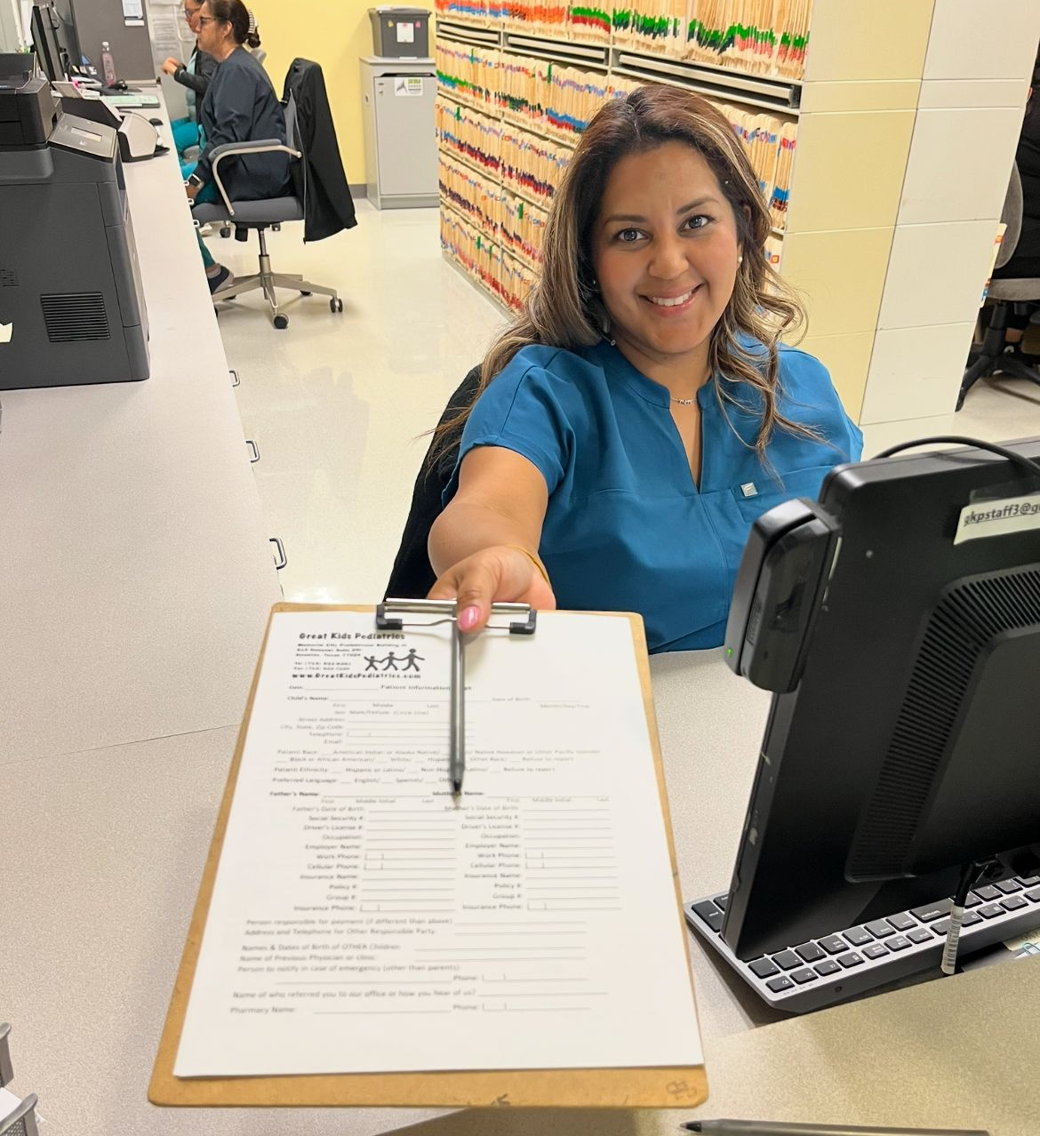 Woman in an office with a computer screen in front of her and medical files in the background.