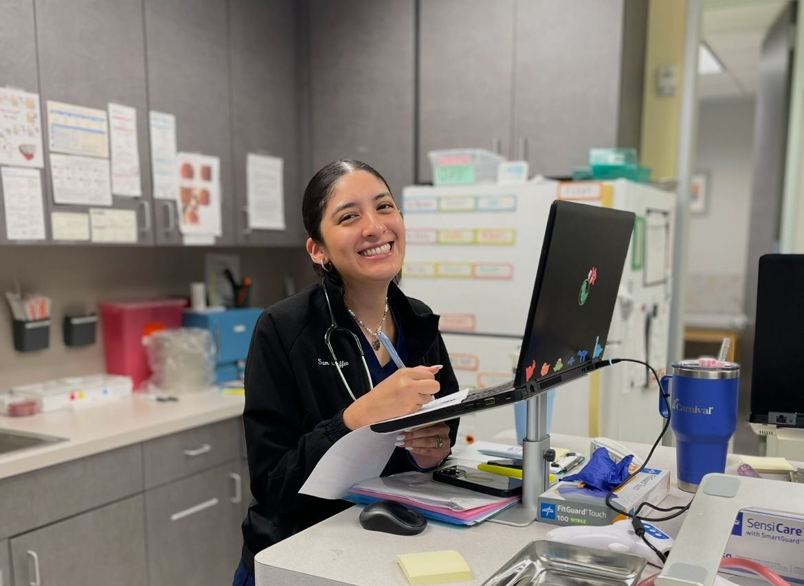 Woman in scrubs smiles while working at a desk with a laptop and papers in a medical setting.