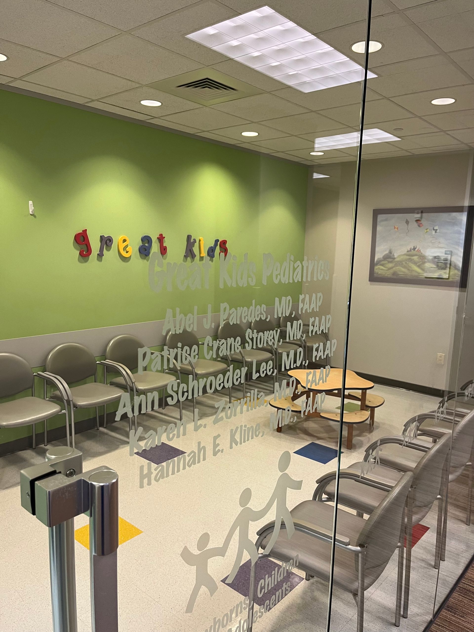 Waiting room of a pediatric clinic with green walls, rows of chairs, and etched glass door with names and logo.