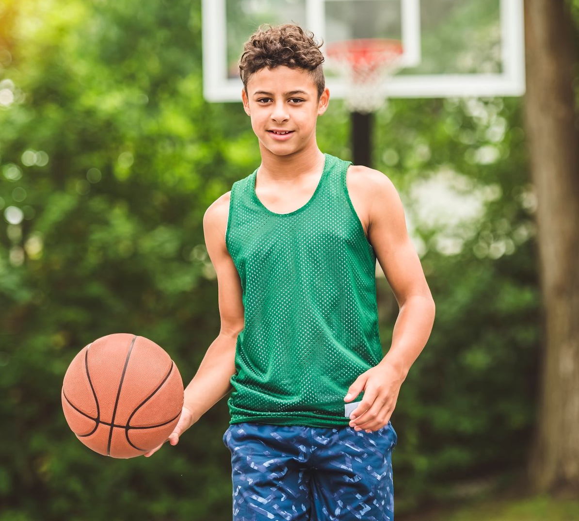 Boy in green jersey holding a basketball near a hoop, smiling outdoors.