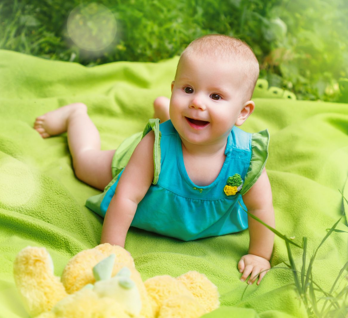 Smiling baby in teal dress on a green blanket on top of green grass.