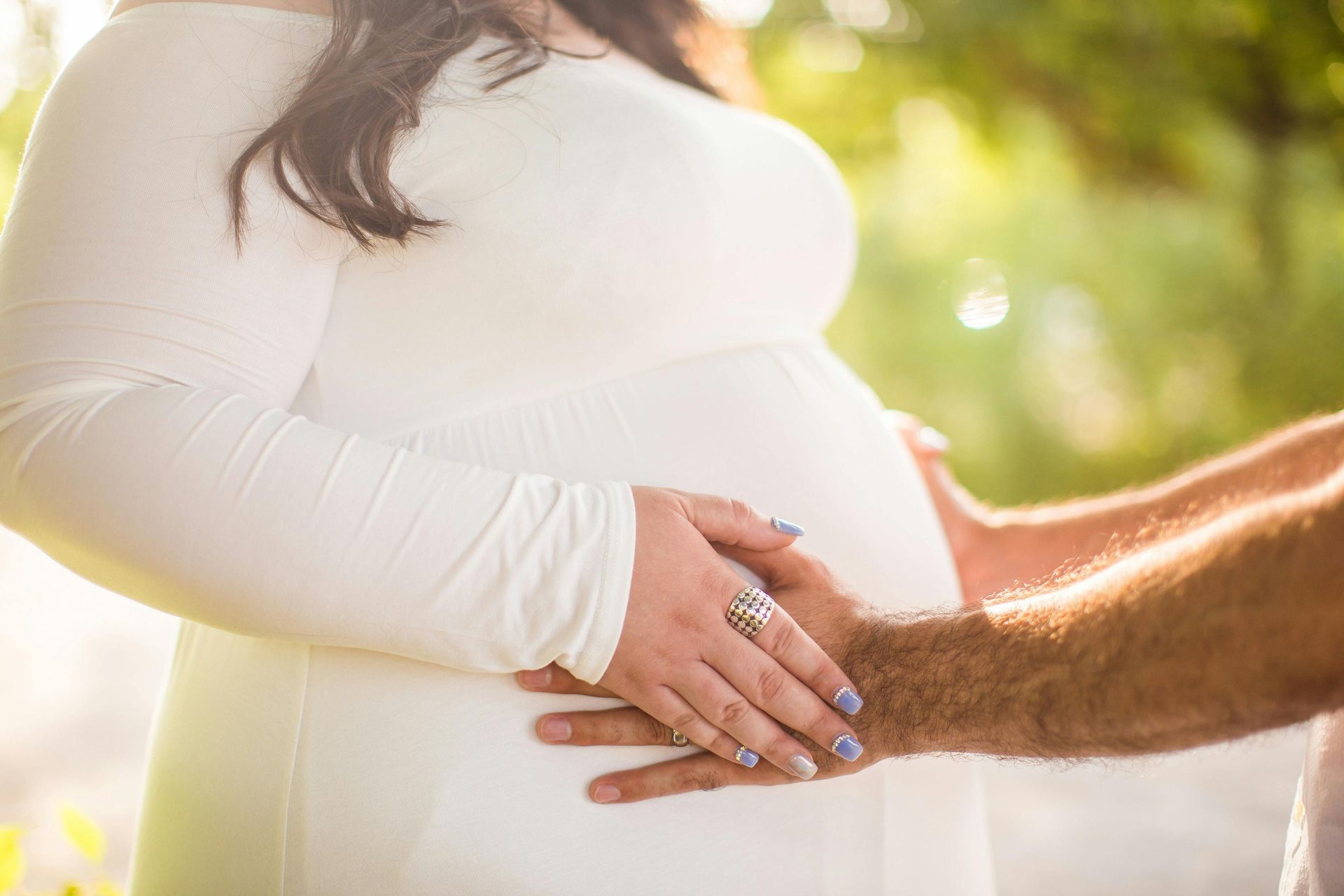 A pregnant woman with trees in the background and hands on her belly.