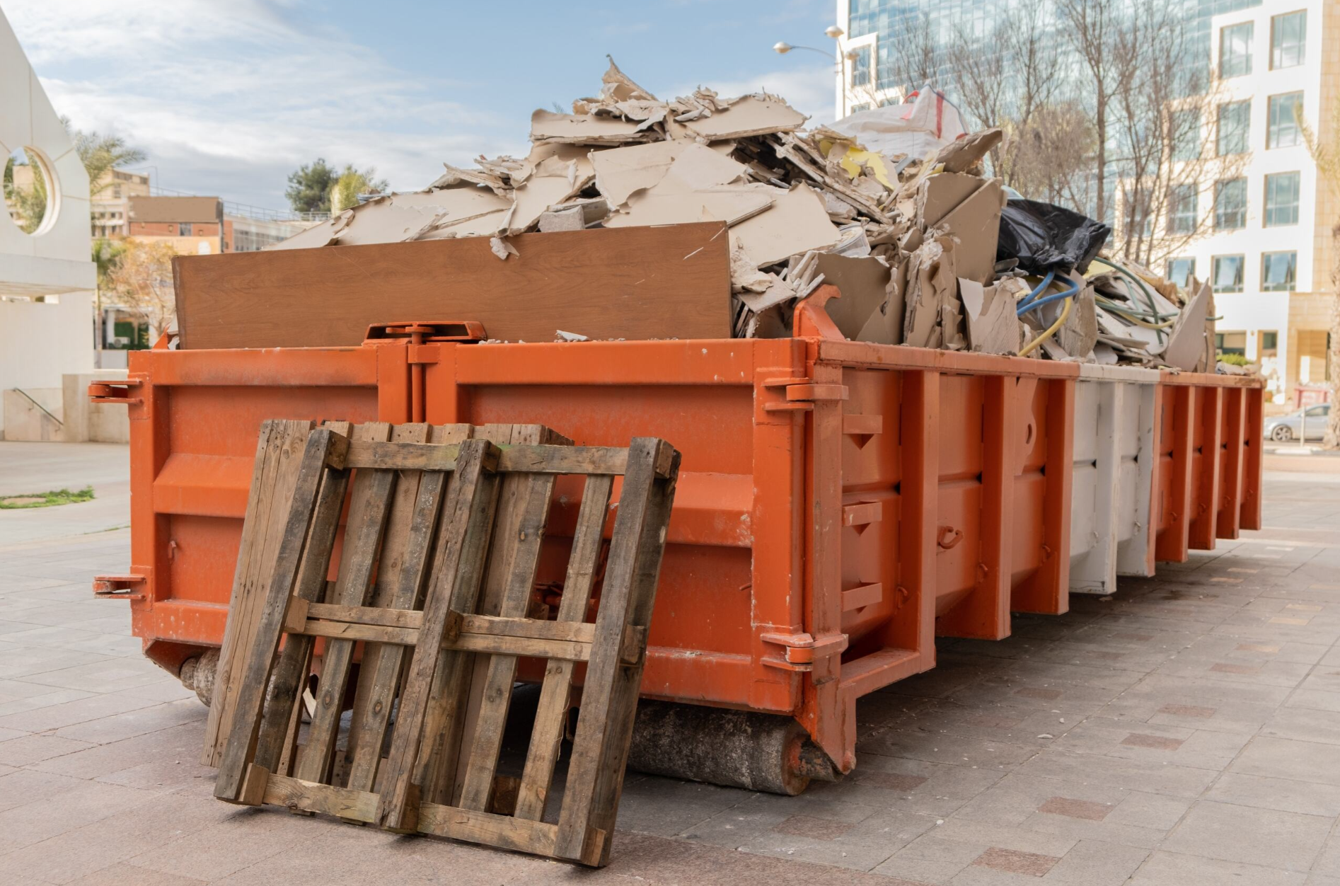 An orange roll-off dumpster filled with construction debris sits on a paved surface, with a wooden pallet leaning against it.