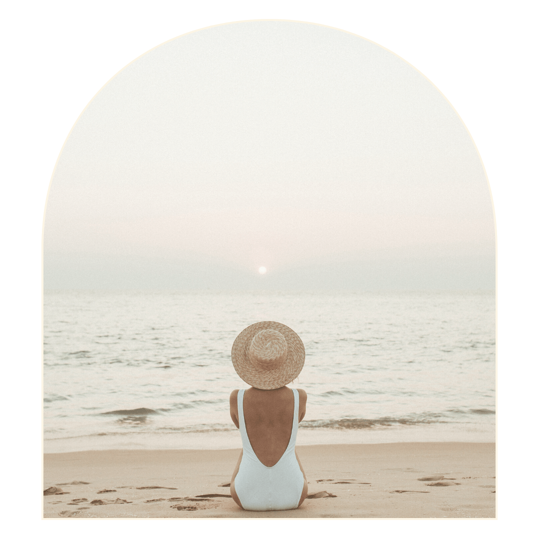 Woman in a white swimsuit and straw hat sits on a beach, facing the ocean at sunset.