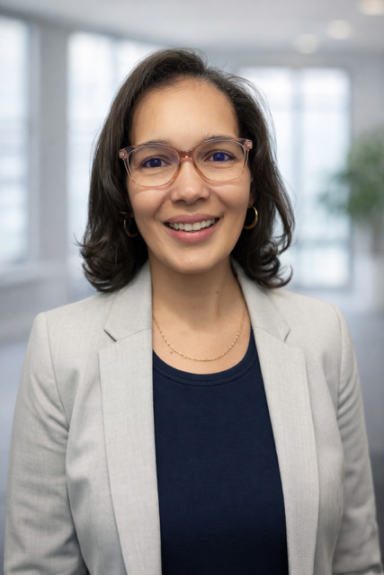 Woman with glasses wearing a light blazer and navy top, smiling. Interior background.