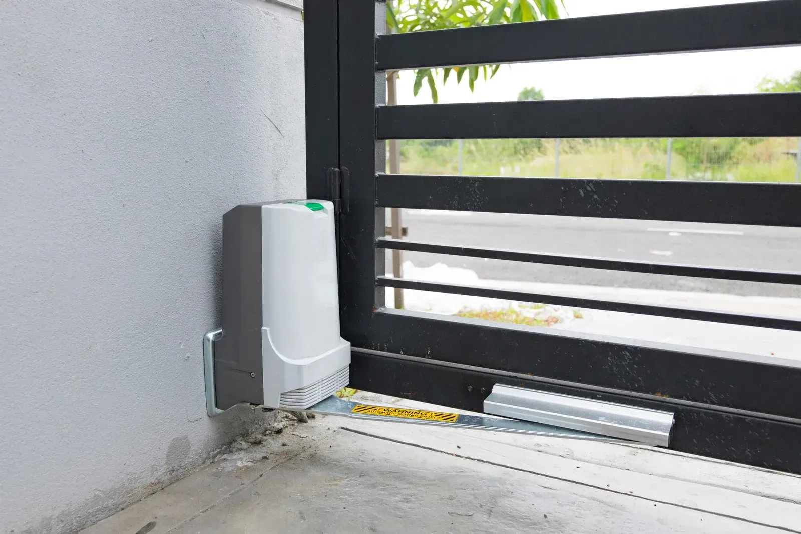 Automatic gate opener attached to a black metal gate and a white wall, outdoors.