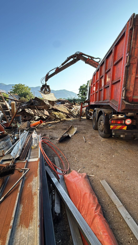 Un camion con un braccio meccanico carica rottami metallici nel suo container sotto un cielo azzurro.