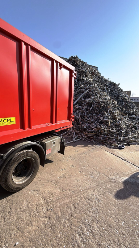 Un camion rosso accanto a un grande mucchio di rottami metallici in una giornata di sole.
