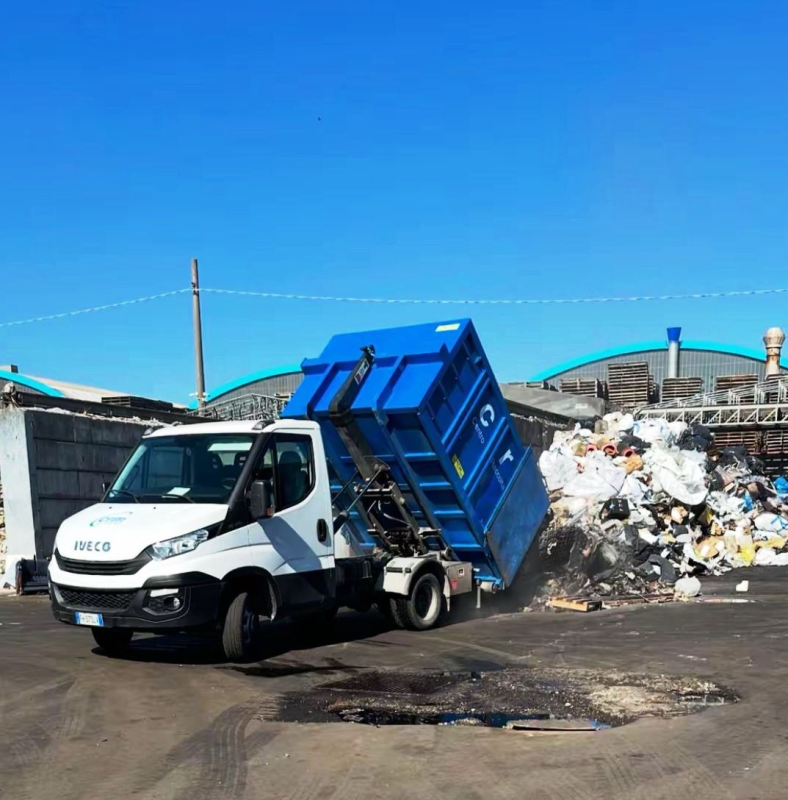 Un camion bianco scarica un contenitore blu di rifiuti in una struttura all'aperto in una giornata di sole.