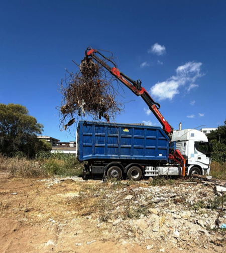Una gru carica materiali di scarto in un cassone blu su un camion contro un cielo azzurro.