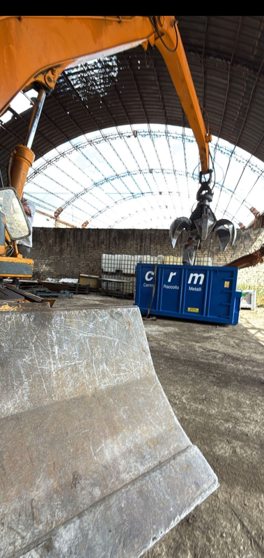 Un braccio di un escavatore che scarica rottami in un cassonetto blu, all'interno di un magazzino.