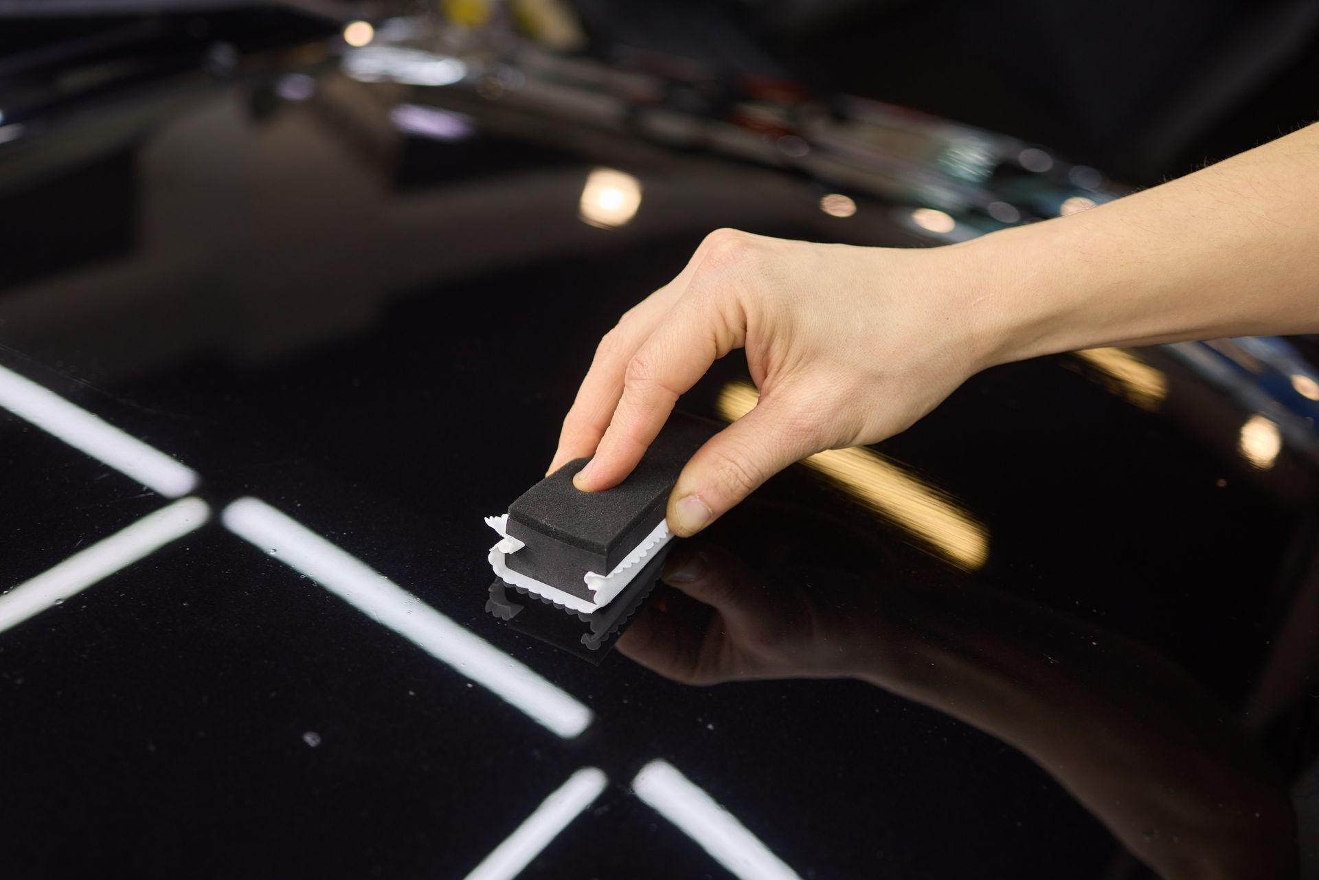 Hand applying coating to a glossy black surface with a black applicator in a brightly lit environment.