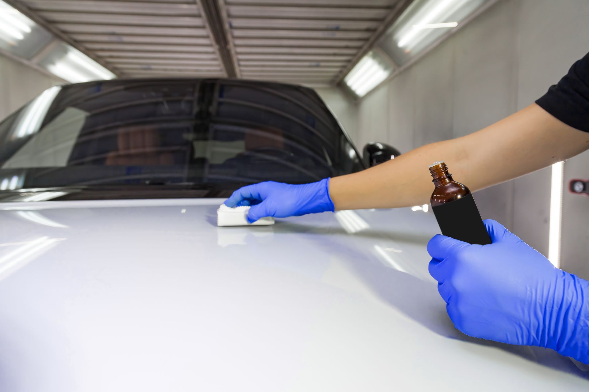 Person in blue gloves applying ceramic coating to a car hood with a bottle in hand.