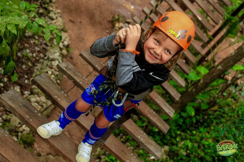 Niño con equipo de seguridad sonriendo en un puente de cuerda de madera en un parque al aire libre.