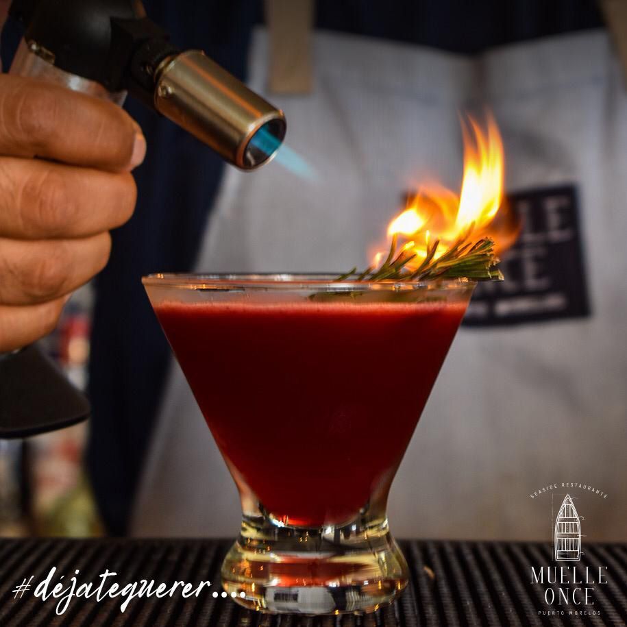 Bartender torching rosemary garnish on a red cocktail in a martini glass.