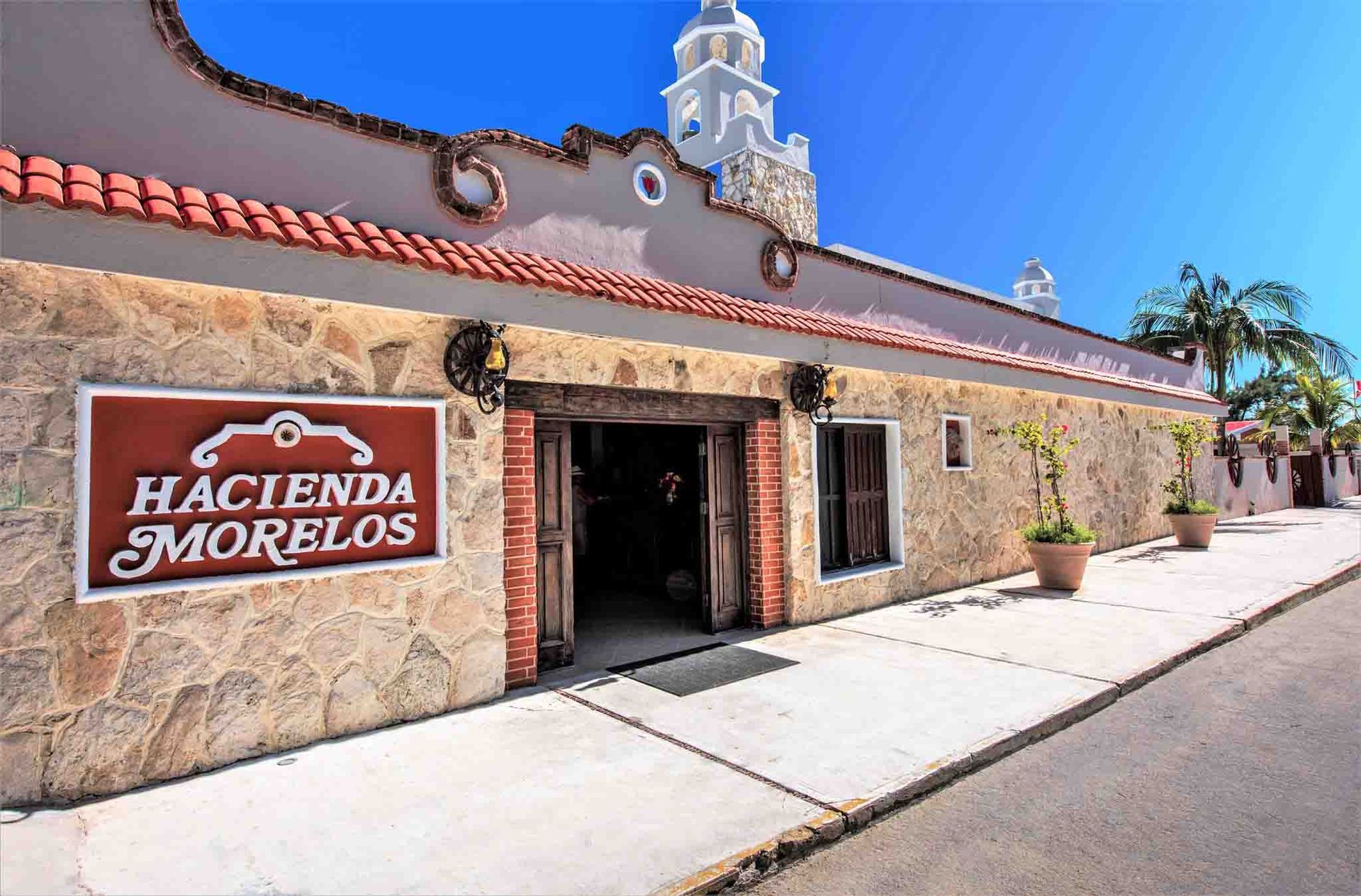 Exterior del restaurante Hacienda Morelos con fachada de piedra, cartel rojo y puerta arqueada bajo un cielo azul.