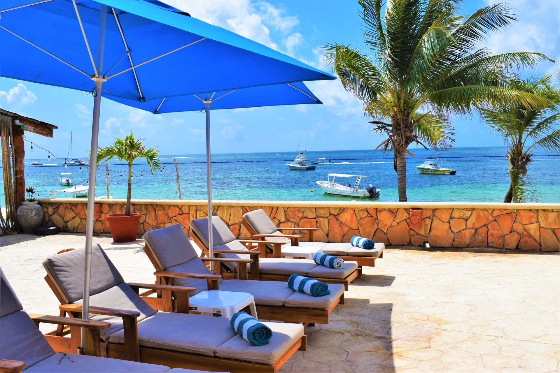 Beach scene with lounge chairs under blue umbrellas, overlooking ocean with boats and palm trees.