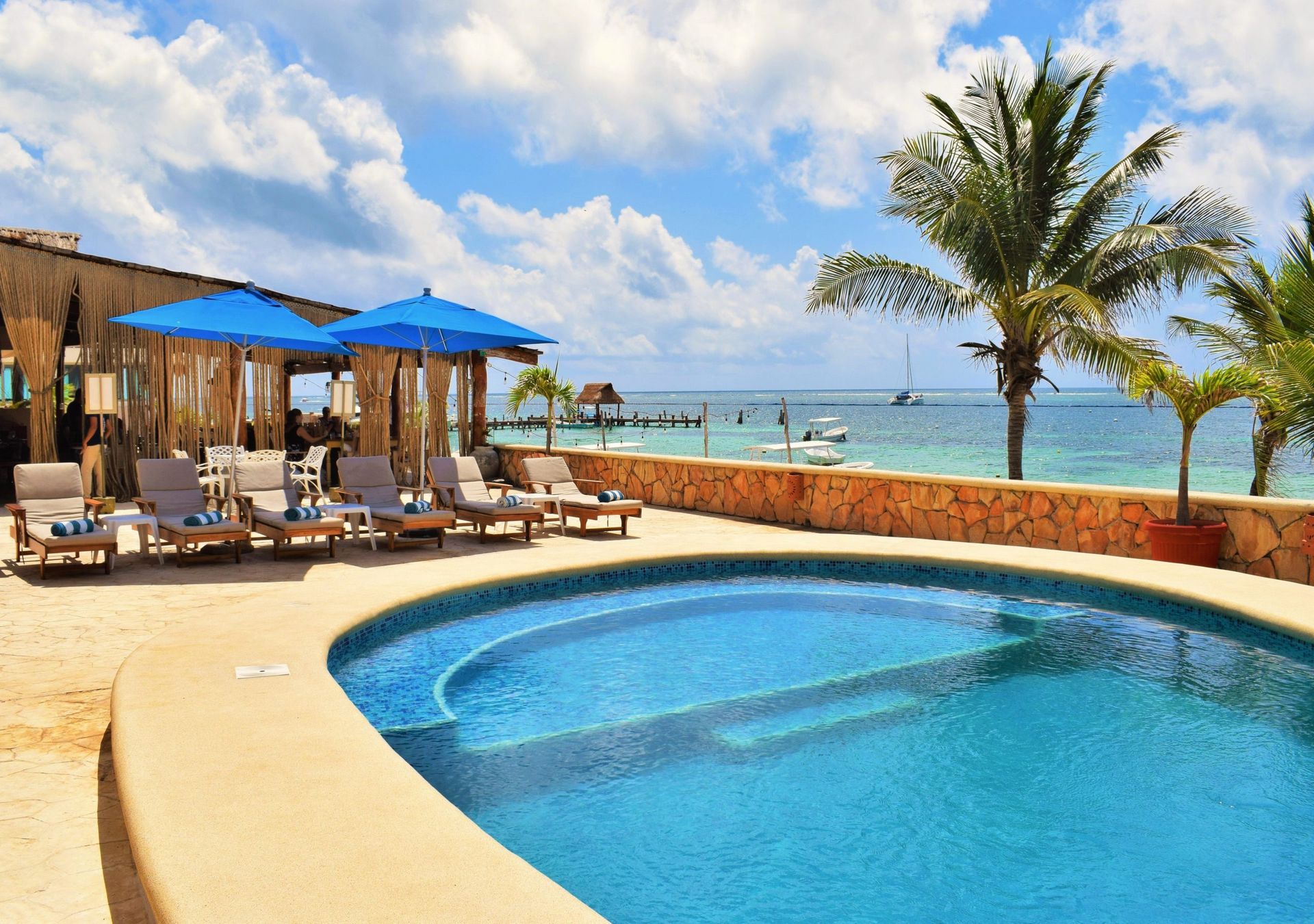 Poolside resort with ocean view; pool, lounge chairs, blue umbrellas, palm trees, and clear blue water.