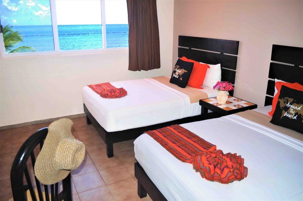 Dormitorio con dos camas individuales y vista al mar. Paredes beige, ropa de cama blanca y detalles en naranja.