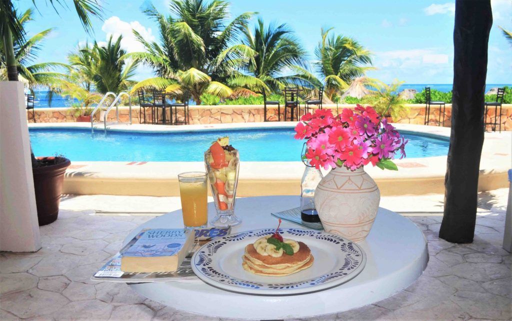 Desayuno junto a la piscina: Panqueques, fruta, flores y jugo en una mesa, con una piscina y palmeras de fondo.