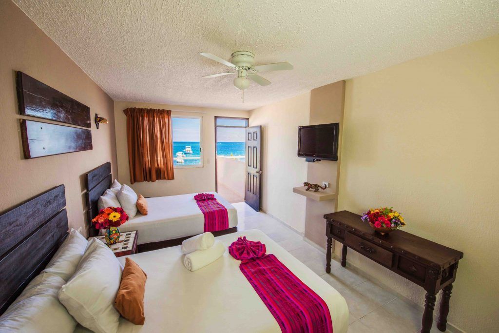 Habitación de hotel con dos camas dobles y vista al mar. Las camas tienen tapetes rosas, TV y una consola de madera.