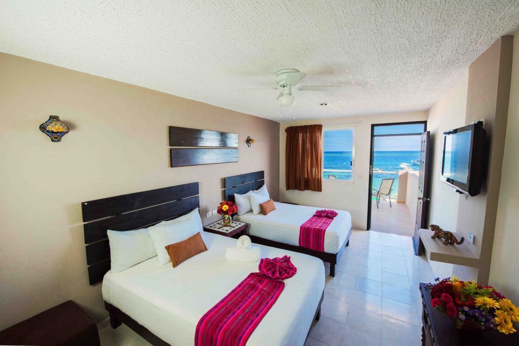 Habitación de hotel con dos camas y vista al mar. Paredes beige, suelo de baldosas, televisión, balcón y decoración floral.