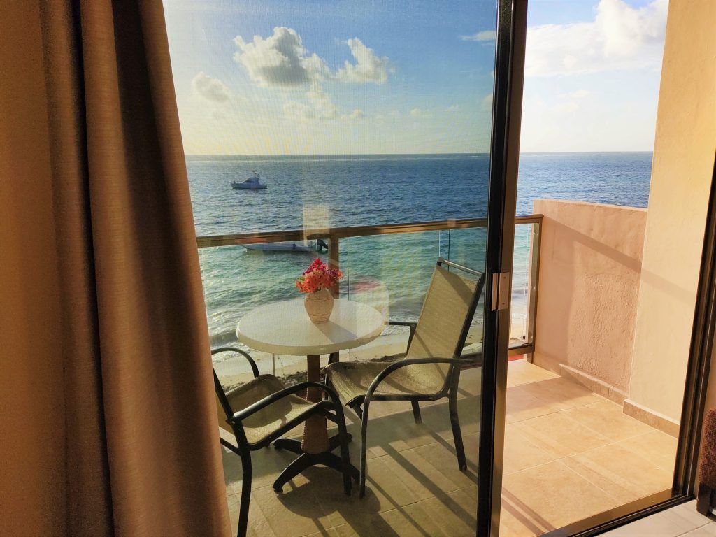 Balcón con vista al mar, con mesa, sillas y flores. Puerta corrediza de vidrio.