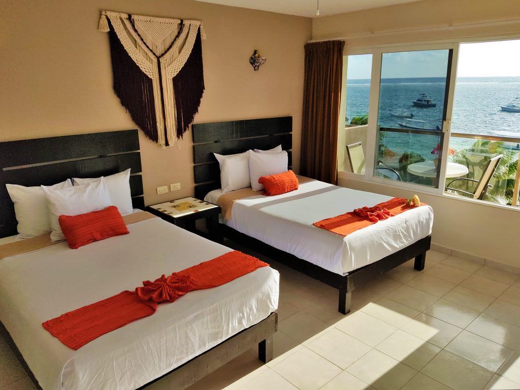 Dos camas en una habitación de hotel con vista al mar. Las camas tienen detalles en naranja; un tapiz cuelga de la pared.