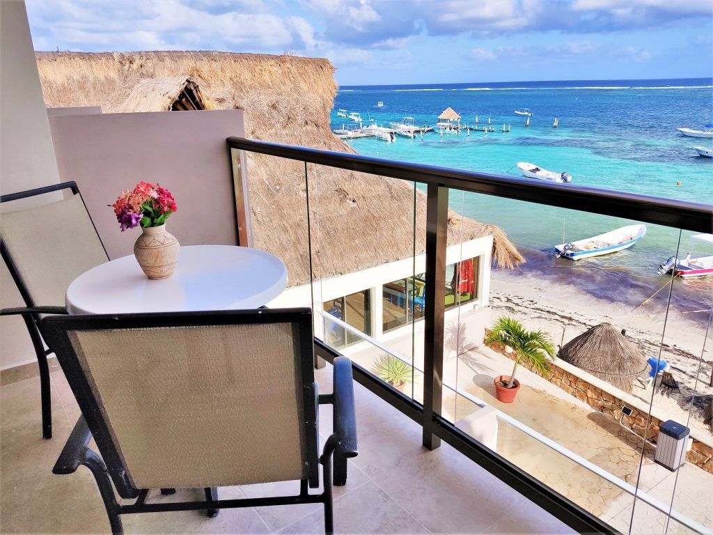 Balcón con vistas al océano turquesa y a la playa. Mesa con flores, dos sillas y barcos.