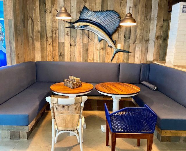 A coastal-themed dining area with a blue sectional, round tables, and a sailfish sculpture on a wood wall.