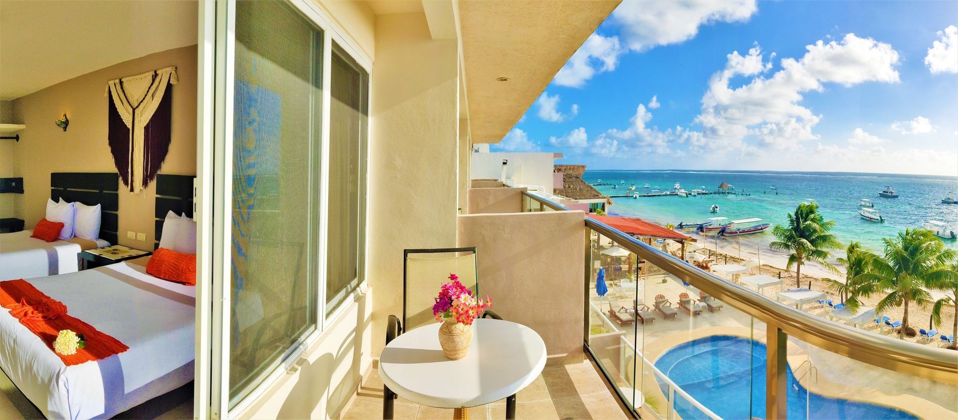 Balcón de la habitación del hotel con vista al mar, mesa, sillas, playa, piscina, cielo azul y nubes.
