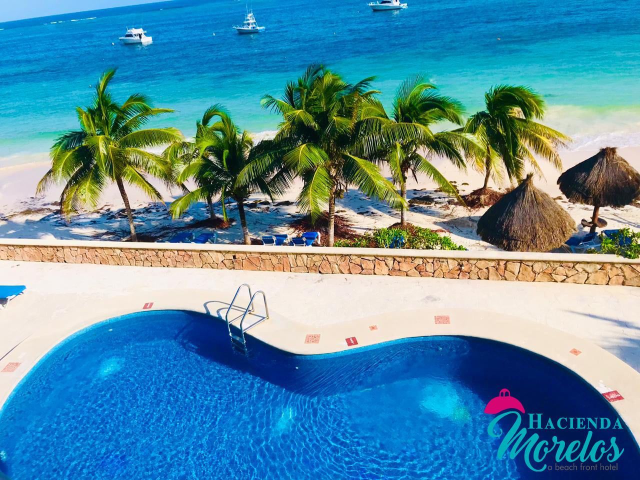 Piscina con vistas a una playa con palmeras y barcos en agua turquesa.