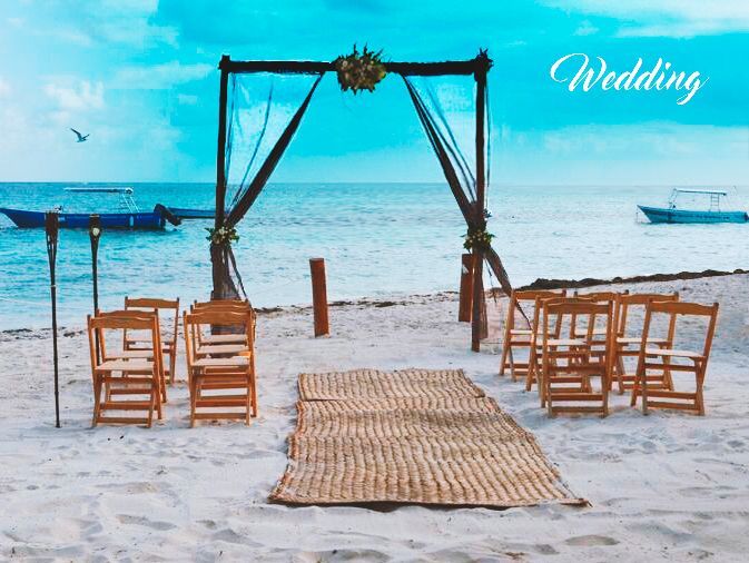 Boda en la playa con arco, sillas y un camino de entrada tejido. Océano y barcos de fondo.
