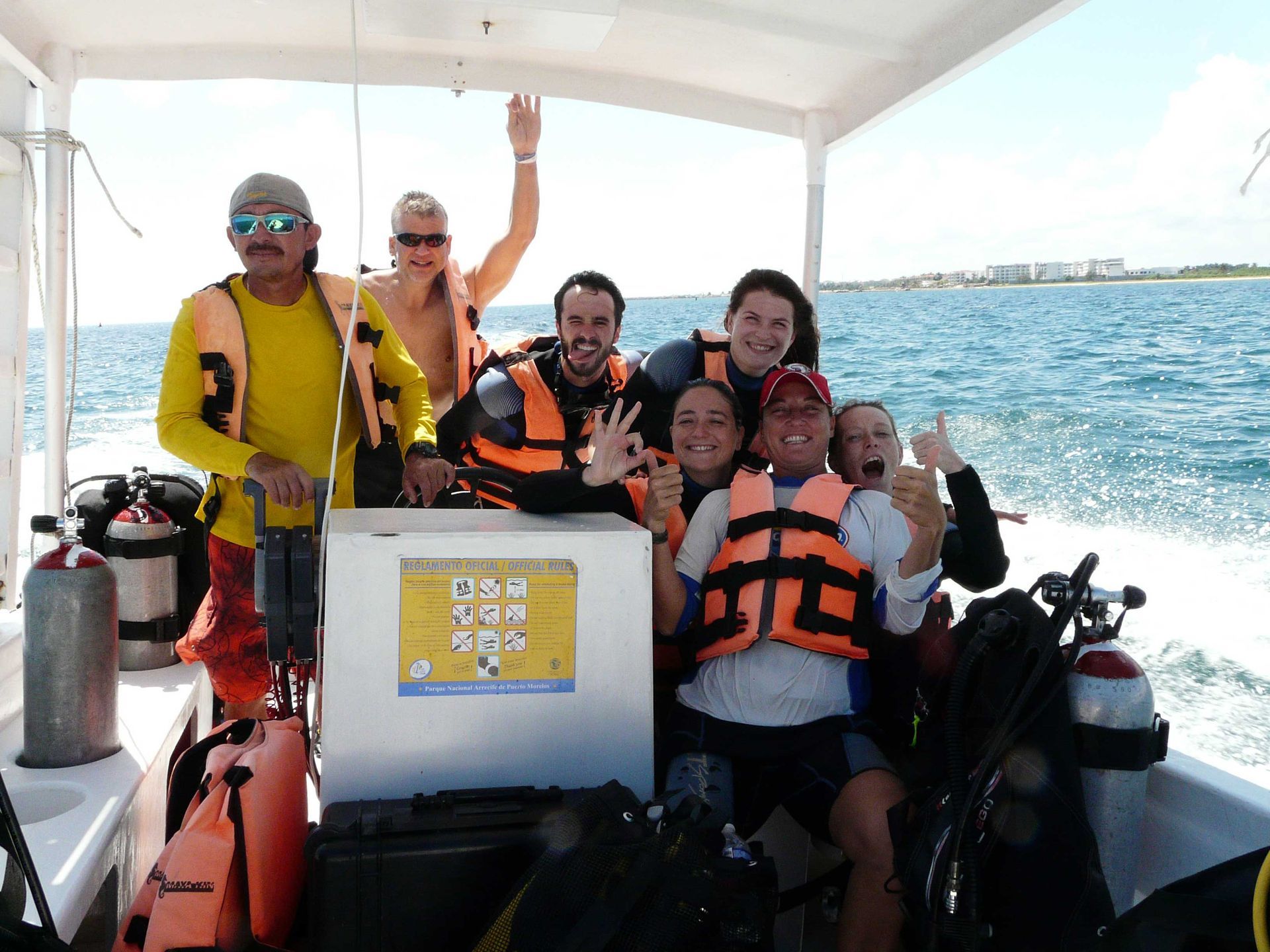 Group of people on a boat, smiling and waving, wearing life vests and scuba gear, on a sunny day.