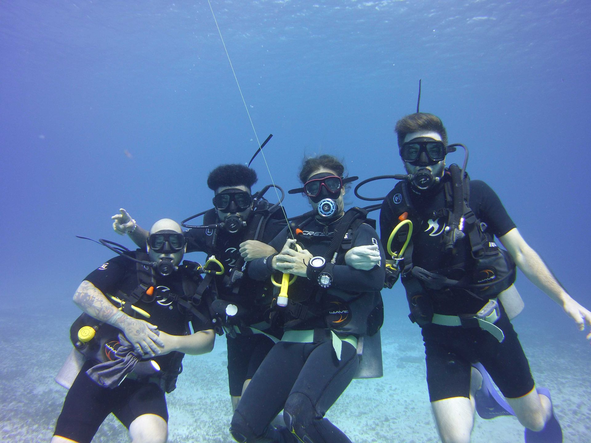 Four scuba divers posing underwater, smiling. They wear black wetsuits, masks, and tanks; blue water and sandy bottom.