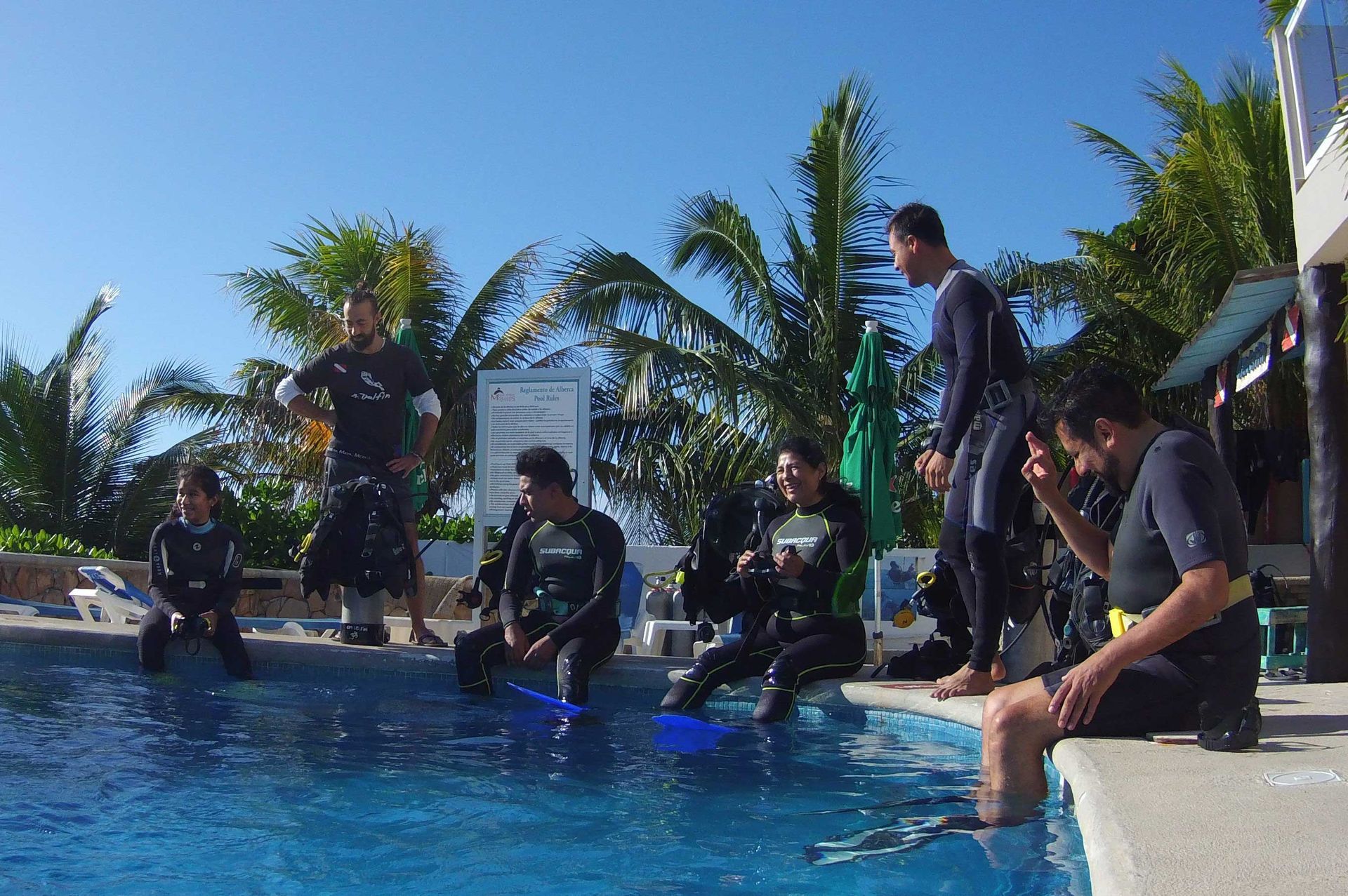 Divers in wetsuits by a pool, preparing to dive. Blue water, sunny day.