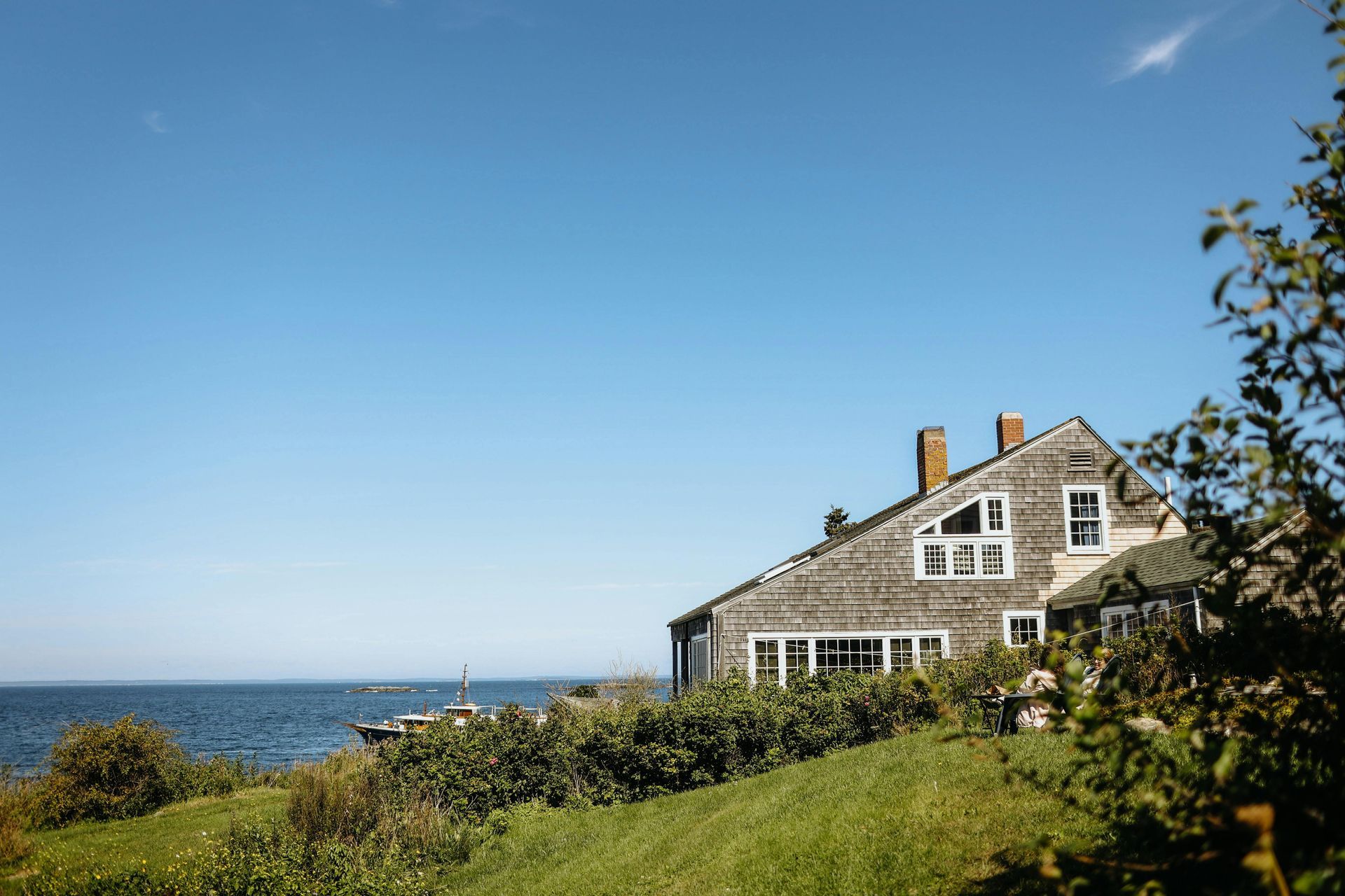 A weathered wooden house with two chimneys sits on a grassy hill overlooking the ocean under a clear blue sky.